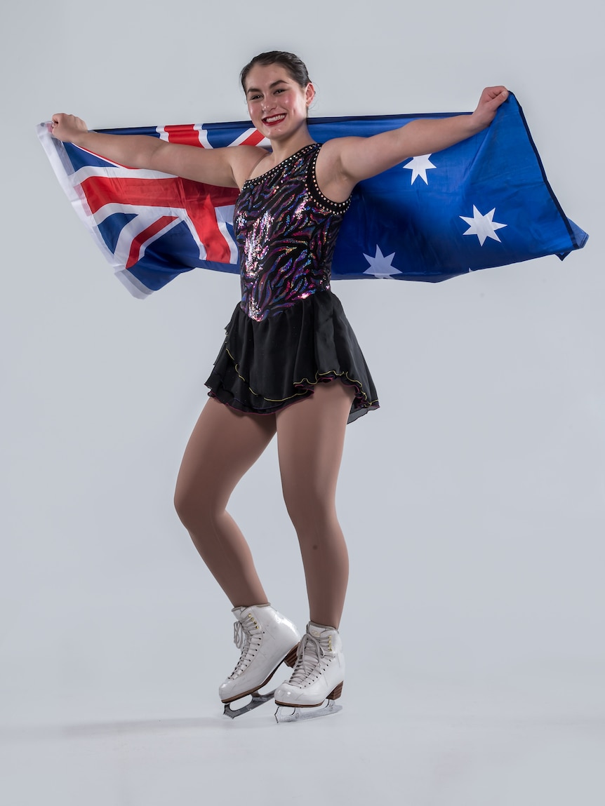 Ada Lacey, a young woman, wears ice skates and a performance leotard, while holding the Australian flag up behind her shoulders.