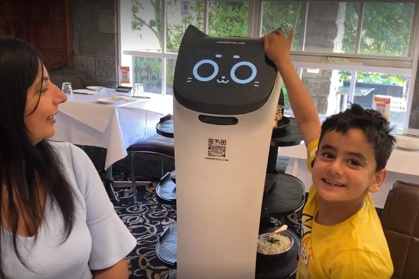 A young boy in a yellow shirt strokes a cat-shaped robot, the cat's digital face is smiling, and there is food on her body.