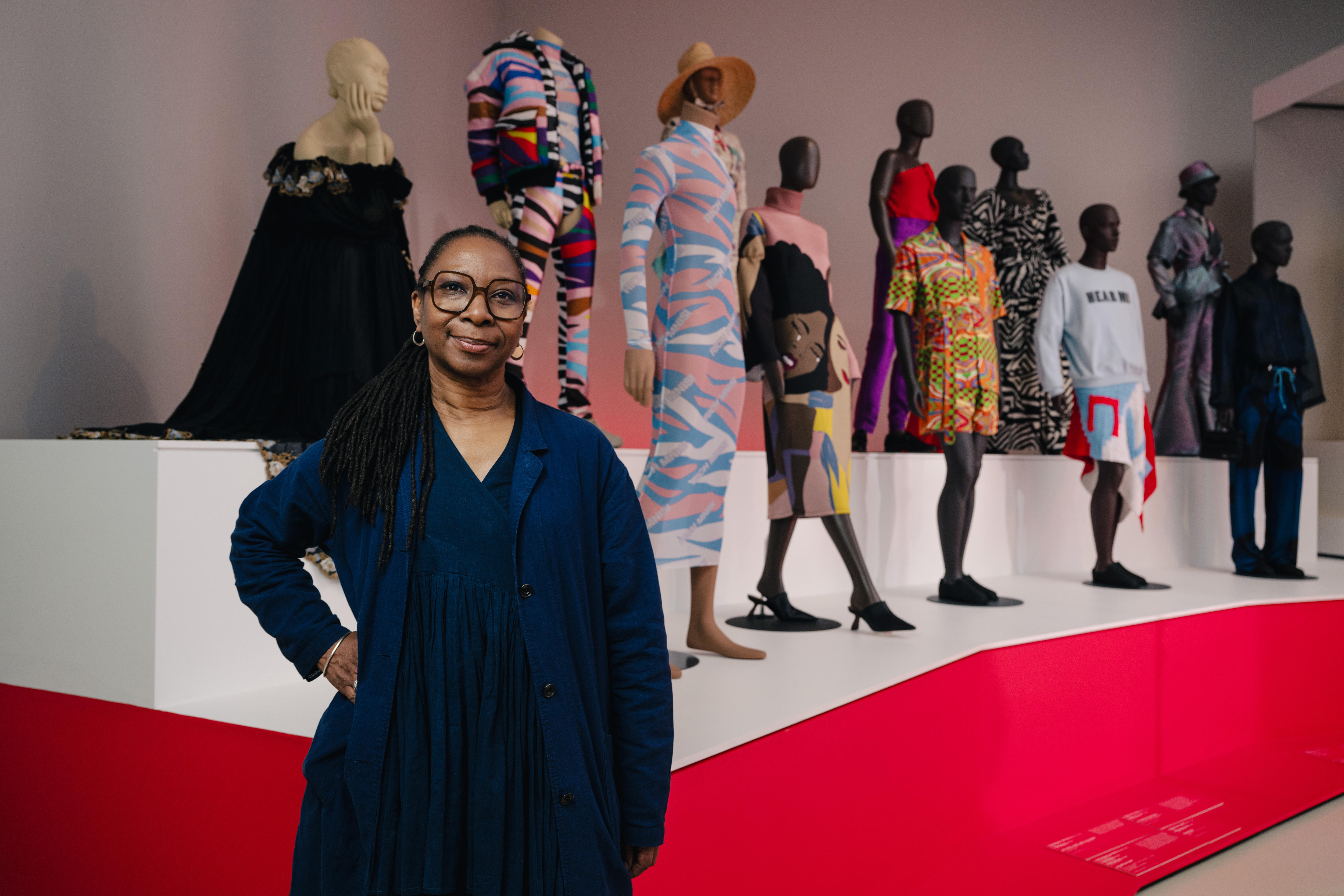 Curator in a blue dress and coat standing in front of mannequins with colourful ready-to-wear designs from African designers. 