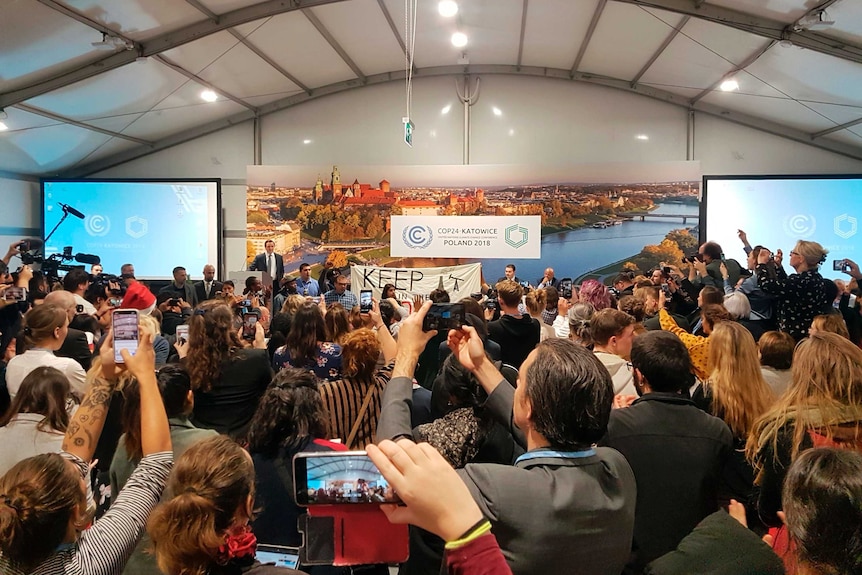 A crowd packed into a mid-sized room hold cameras and phones, recording someone speaking against a COP-24 banner.
