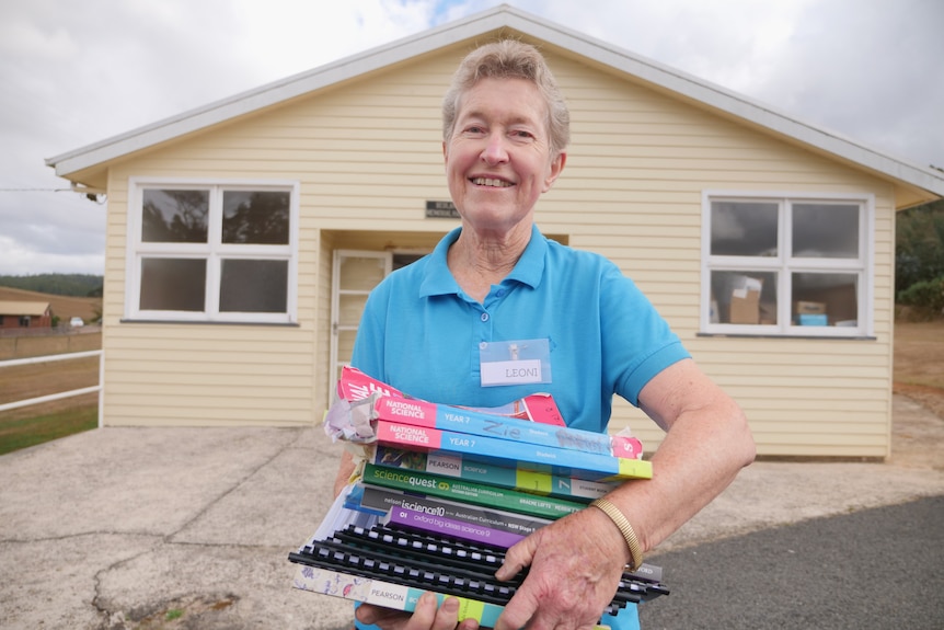 A woman wearing a blue button up shirt holds a pile of books while standing in front of a house.