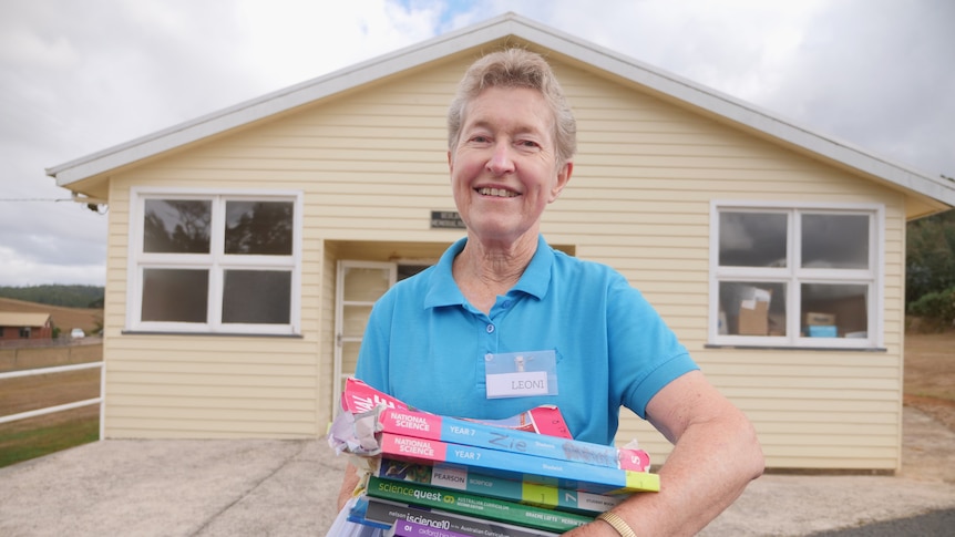 A woman wearing a blue button up shirt holds a pile of books while standing in front of a house.