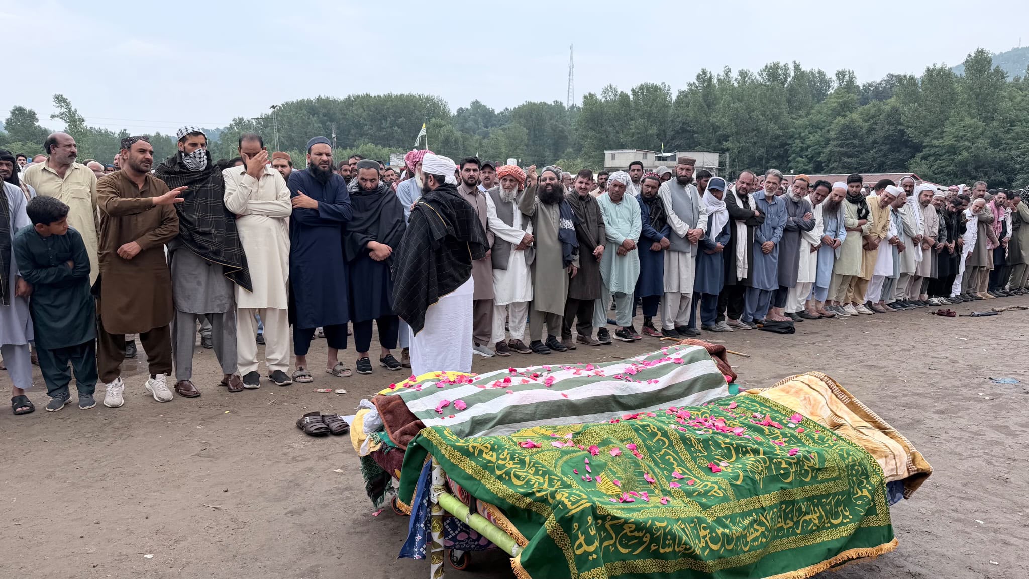 A large group of men stand vigil beside two covered bodies