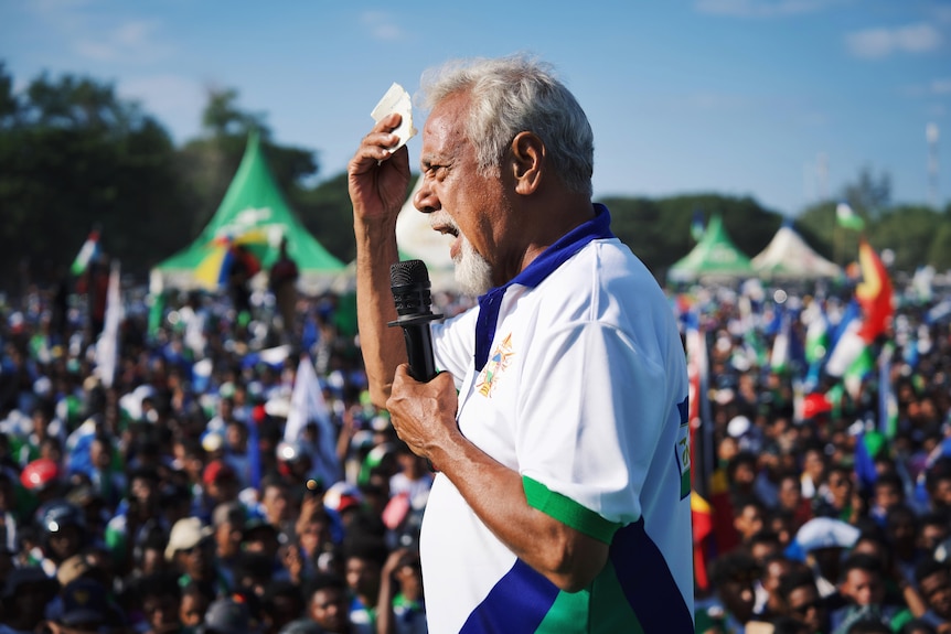 Xanana Gusmao, with white hair raising one hand in air while speaking into microphone, rallies the crowd
