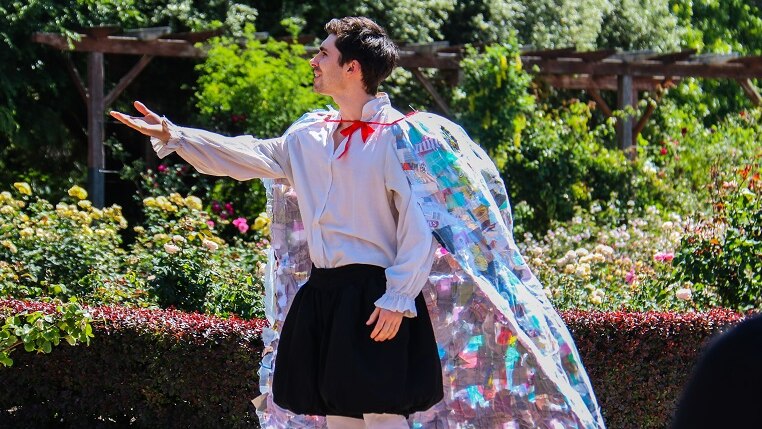 An actor performs Shakespeare in St Kilda Botanical Gardens wearing a cape made from trash on a sunny day.