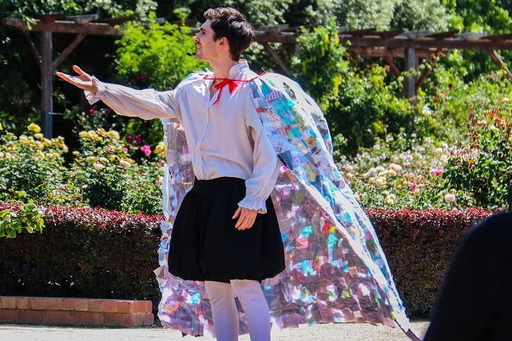 An actor performs Shakespeare in St Kilda Botanical Gardens wearing a cape made from trash on a sunny day.