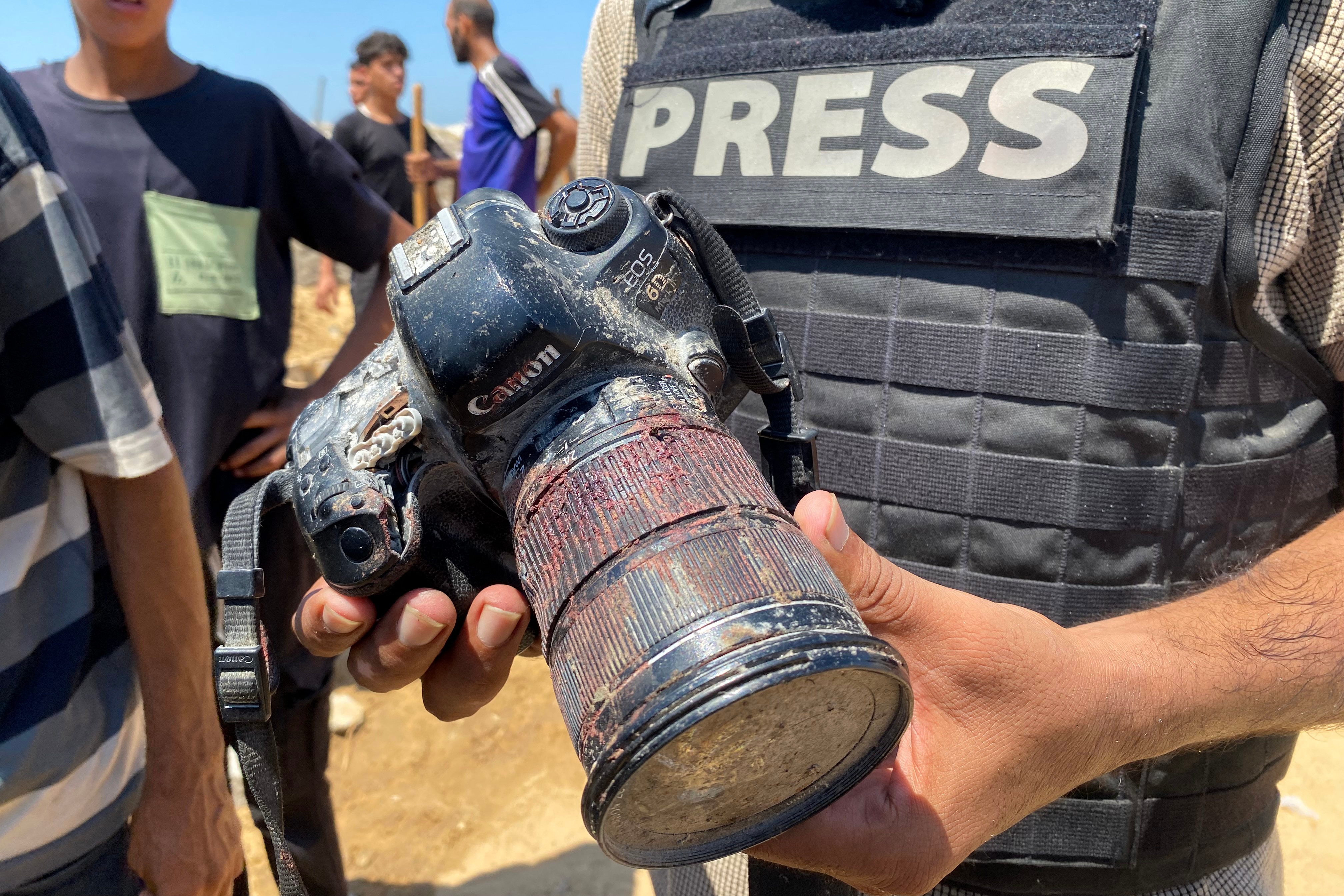 A journalist wearing a press vest holds a camera that is covered with dust and blood