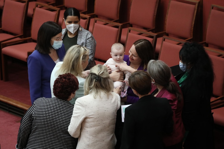 Green stands on the senate floor holding her baby, surrounded by a group of female politicians giving it attention.