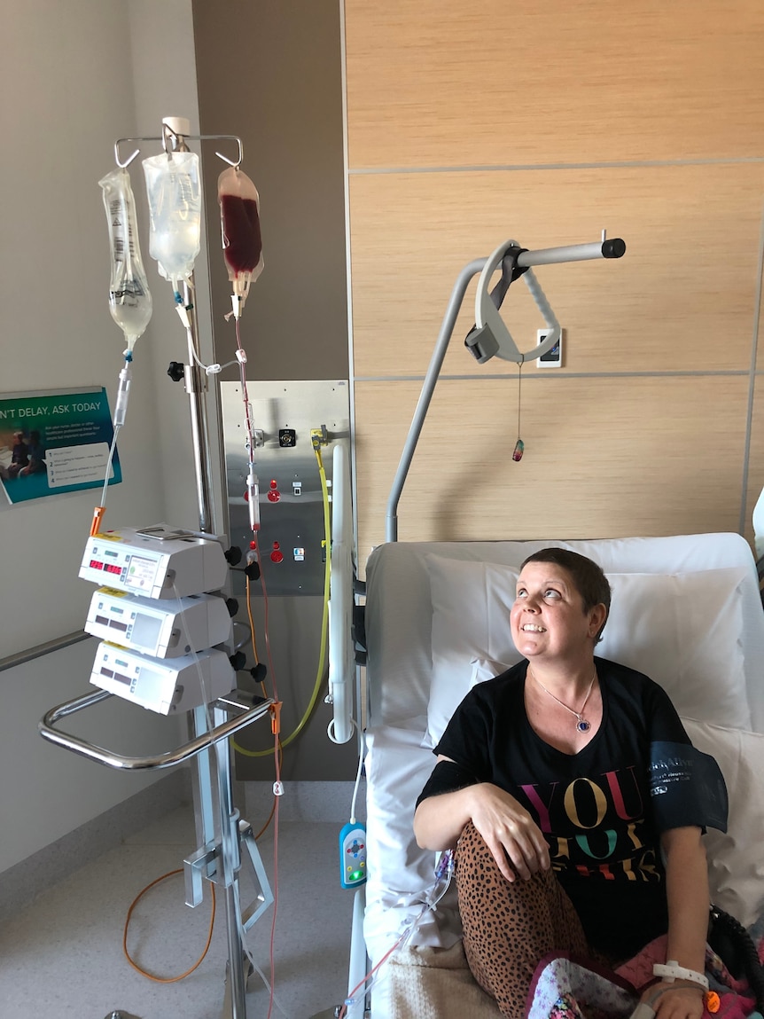 A woman sits in a chair in a hospital room with a bag of blood stem cells and fluids infusing from a cart next to her.