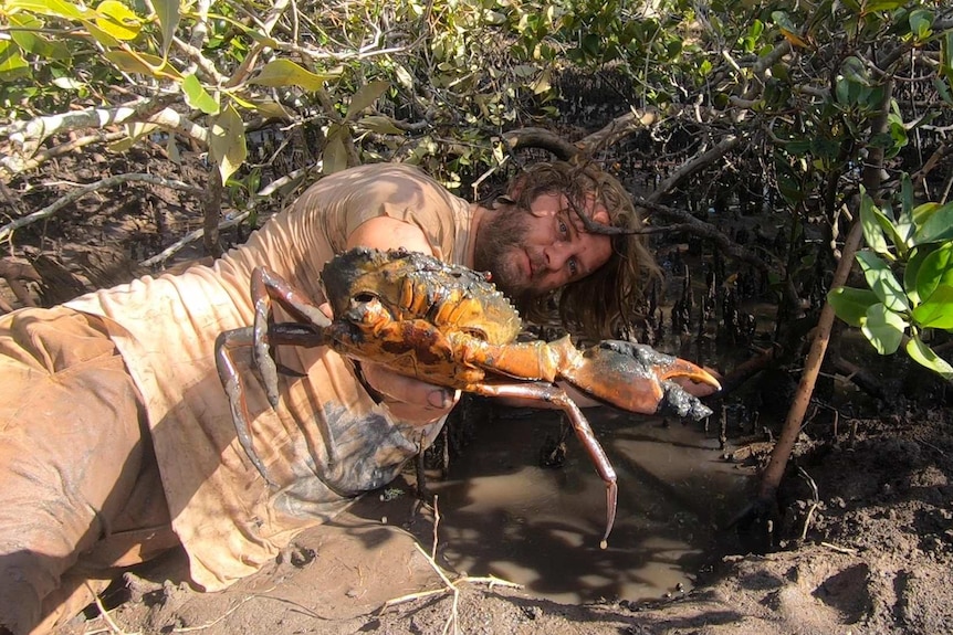 A man lying in mud with a crab in his hand.