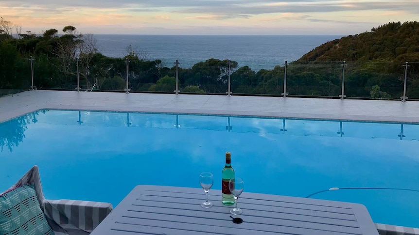 A deck with a pool at sunset. A bottle of wine with two glasses in the foreground.