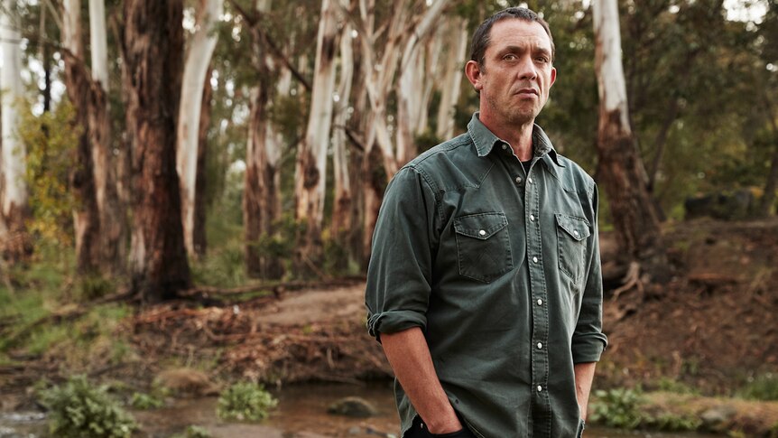 Angus Cerini stands hands in pockets outdoors in bush, with snowgums behind him, staring at camera, looking serious.