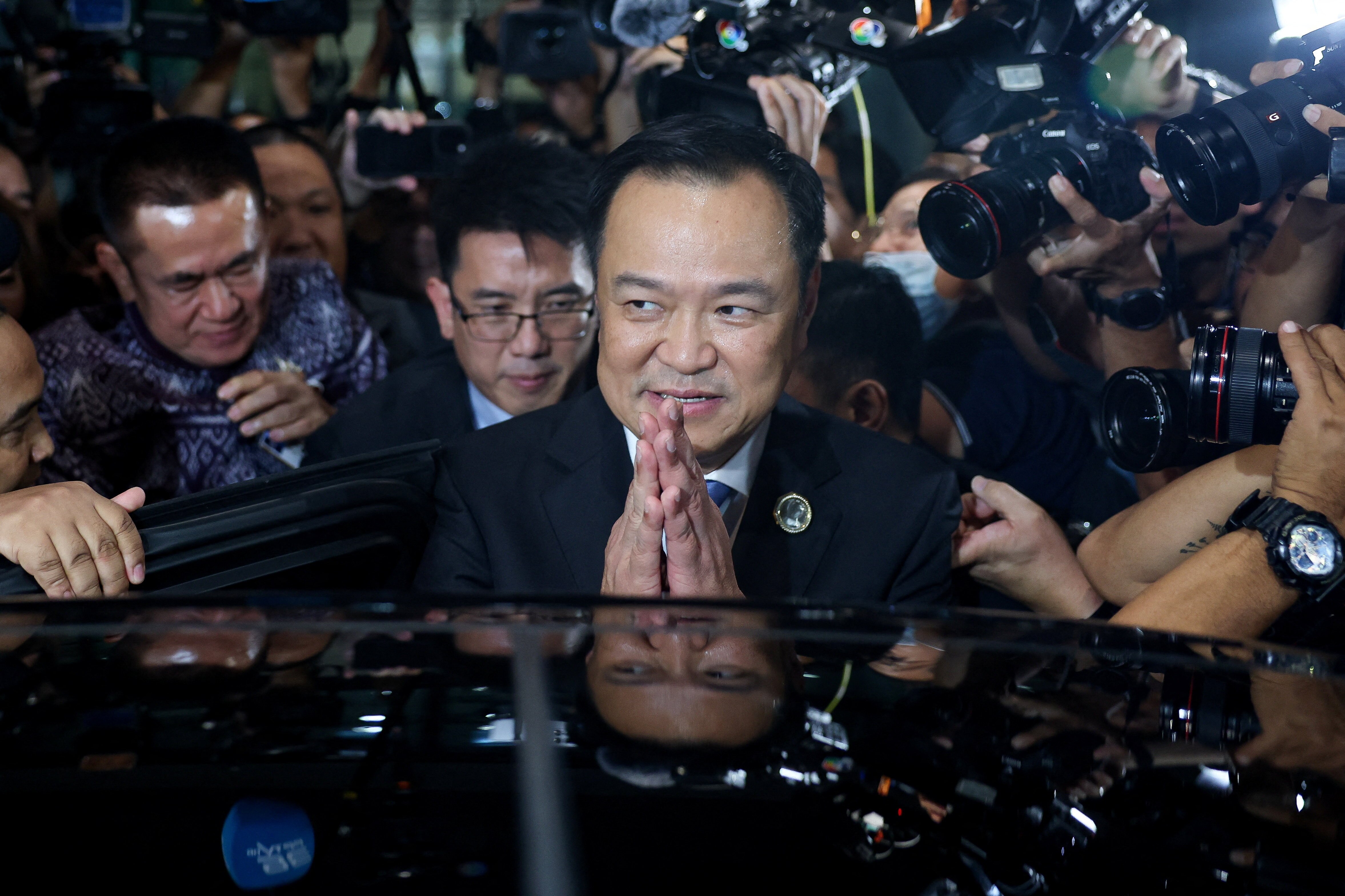 A man clasps his hands in a gesture of thanks while getting into a black car which is surrounded by people and cameras