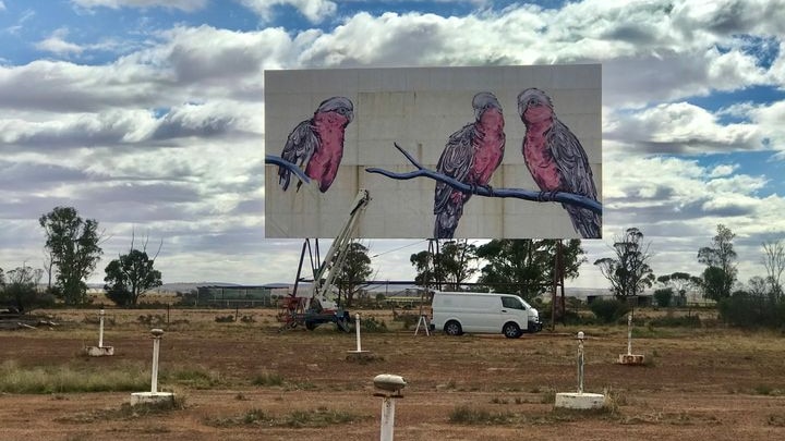 An old drive in movie screen in the distance is painted with large birds, a van and old speakers sit in front. 