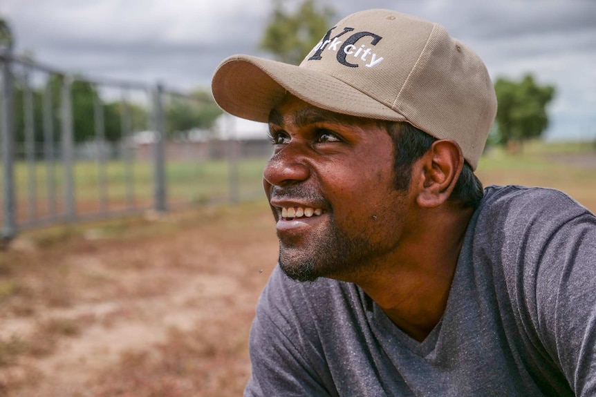 A young indigenous man looks out of frame to the left. He wears a cap, and a fly has landed on his unshaved cheek.
