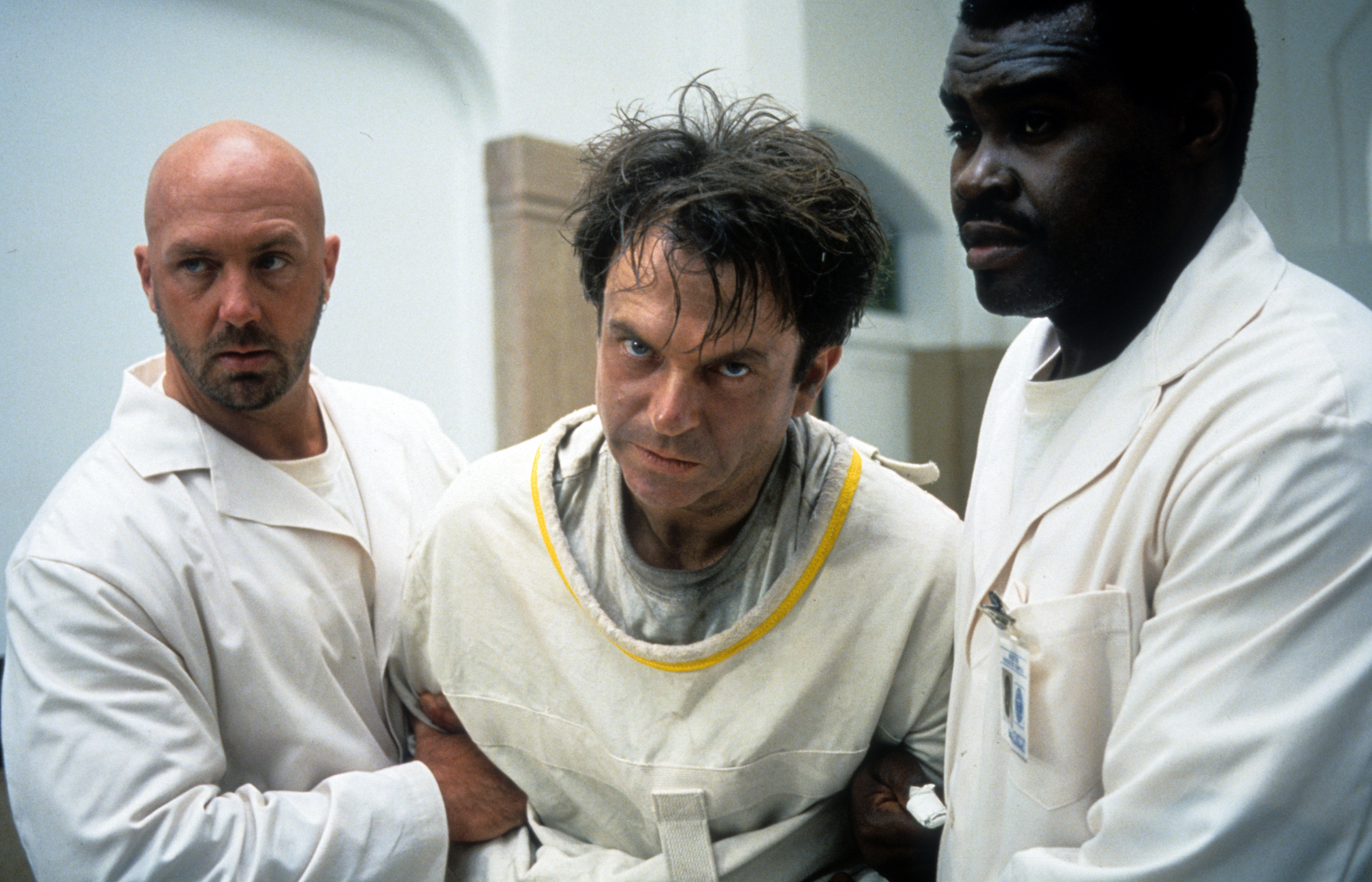 Movie still showing Sam Neill in a medical white gown being held up by two men in white medical jackets