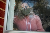A woman in a pink jumper and face mask stares out the window of her home.