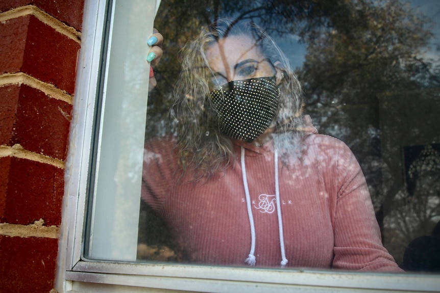 A woman in a pink jumper and face mask stares out the window of her home.