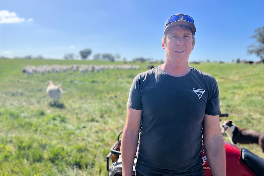 Drew Symons wears a dark t-shift, cap and sunglasses rests on hat. Goats and dog in background.