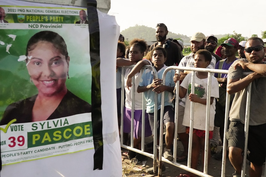 A sign displaying a woman smiling with Pascoe written below it as a crowd of people look on from behind a railing.