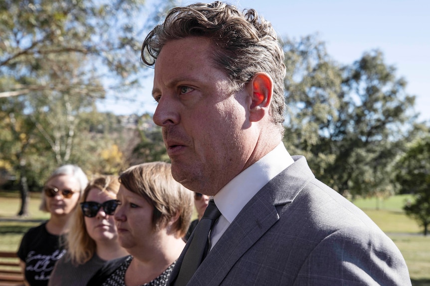 A man in a grey suit stands in front of journalists.