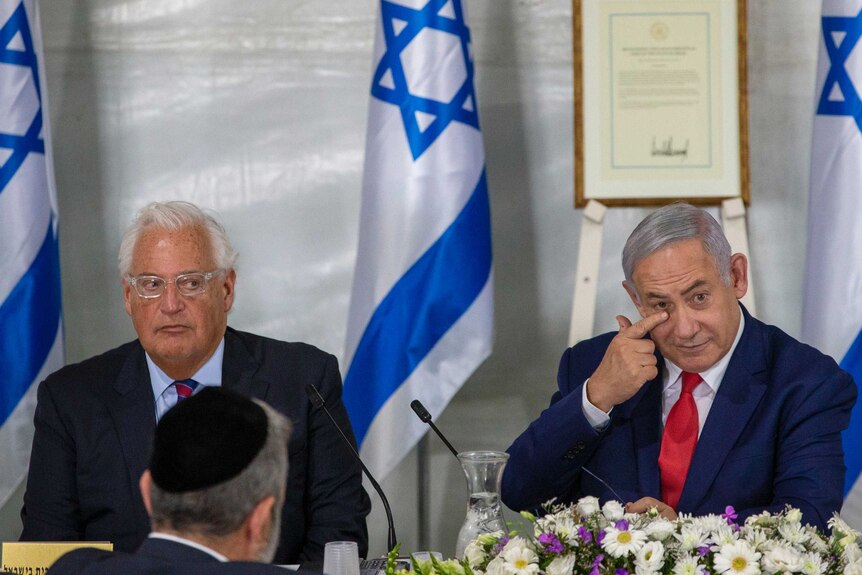 David Friedman and Benjamin Netanyahu sit next to each other at a table dressed with flowers, Israeli flags in the background.