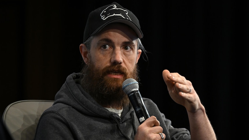 A man wearing a black cap with a beard speaks into a microphone.