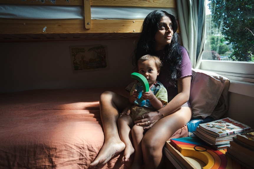 A woman holding her bub and sitting on a bed, looking out a window.