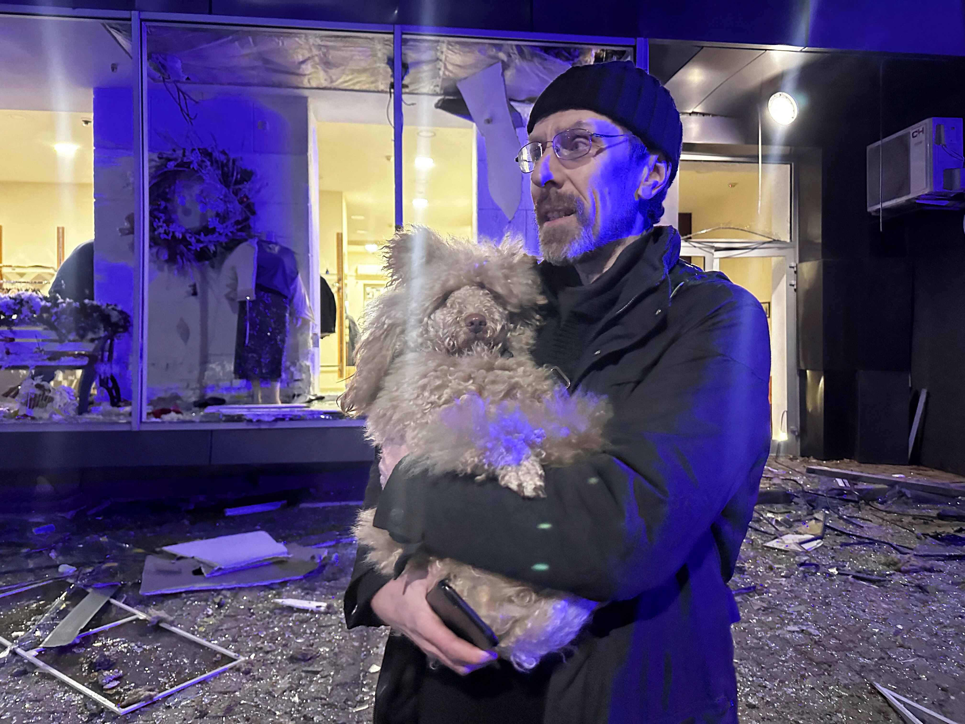 A man holds a dog in front of a damaged building.