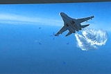 A screenshot of a video showing a fighter jet sailing through the air with clouds behind it. 