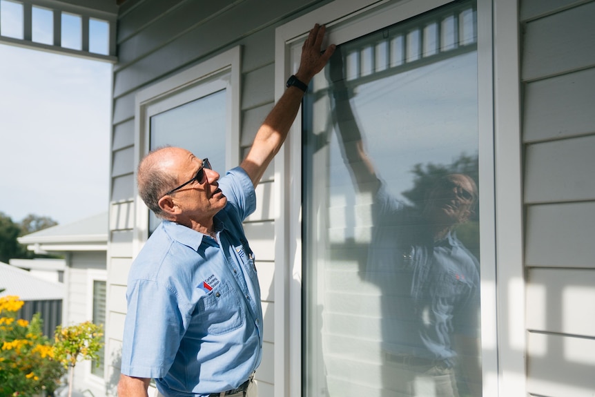 A balding man wearing dark sunglasses and a work shirt holds his hand up against a window frame on a house.