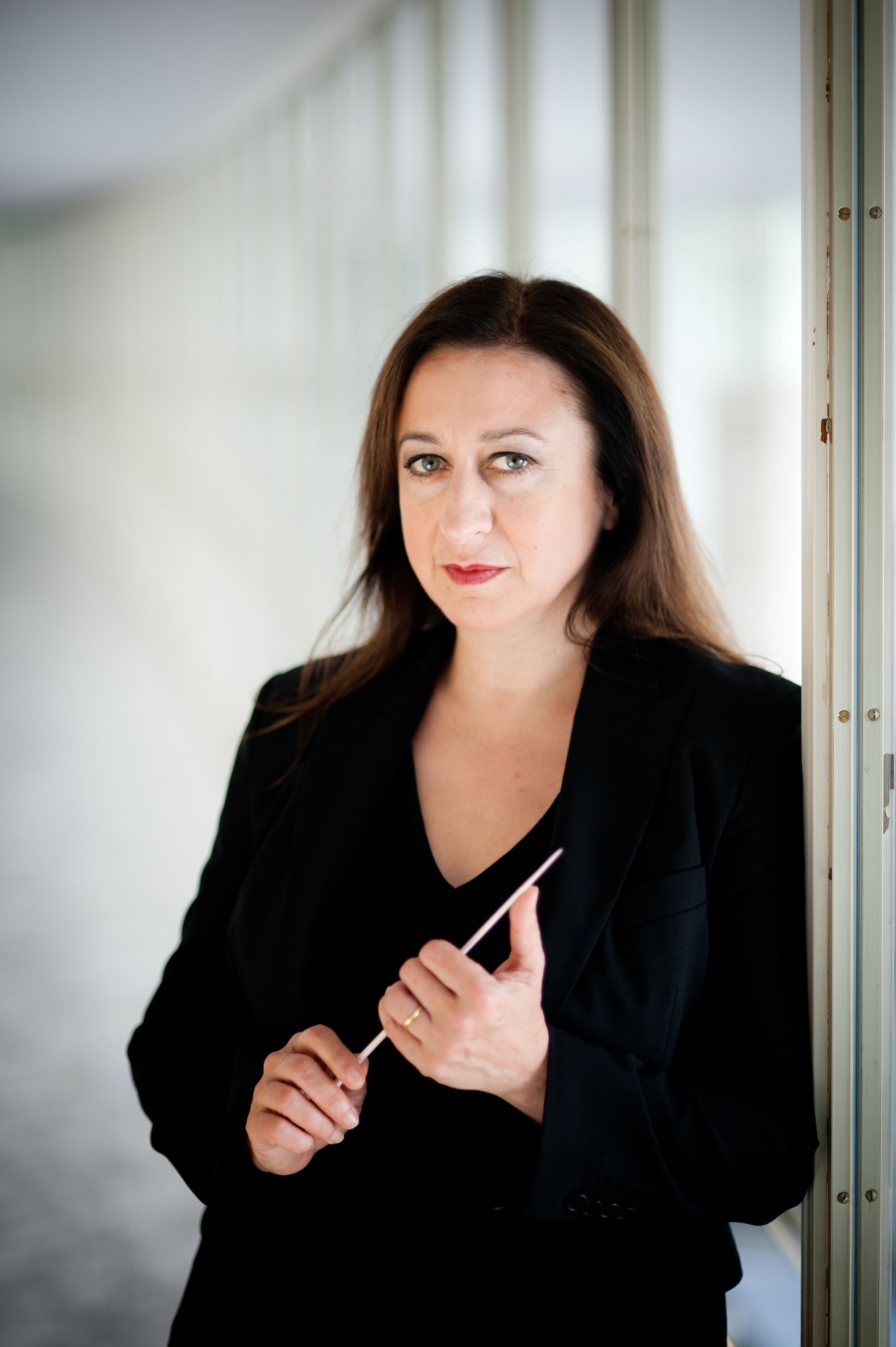 White middle-aged woman with long dark hair wears a black suit and leans against a wall in a white, brightly-lit foyer.