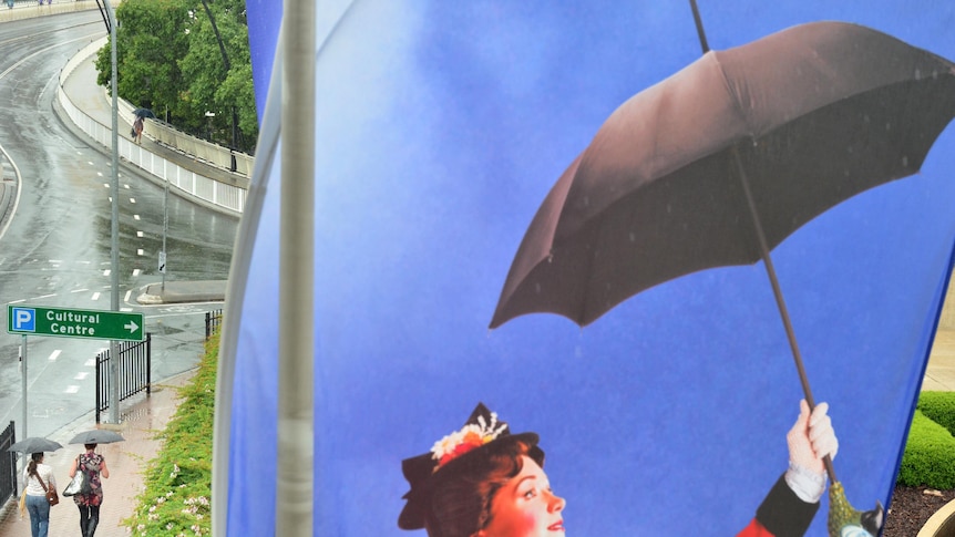 Two people under umbrellas walk past a Mary Poppins banner
