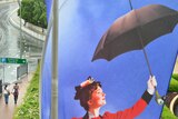 Two people under umbrellas walk past a Mary Poppins banner