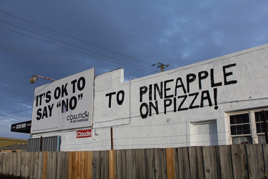 A handpainted message next to a billboard on a pizza shop wall.