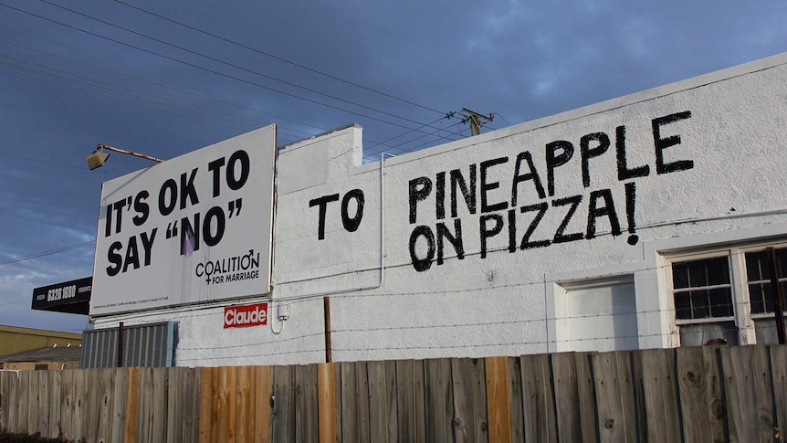 A handpainted message next to a billboard on a pizza shop wall.