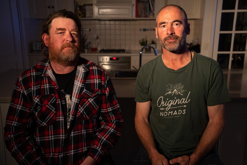 Two men sitting in front of a kitchen. The man on the left wears a plaid shirt and the man on the right a green t-shirt.