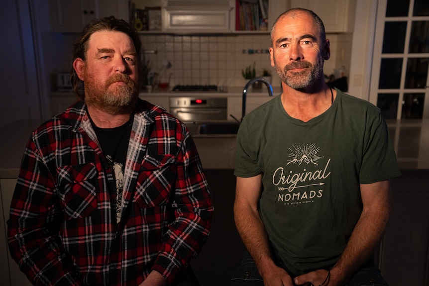 Two men sitting in front of a kitchen. The man on the left wears a plaid shirt and the man on the right a green t-shirt.