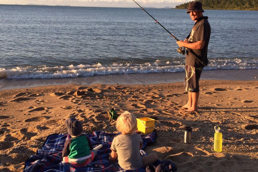 Grant McEwan smiles as he fishes on a beach, looking at his two children sitting on a blanket on the sand.