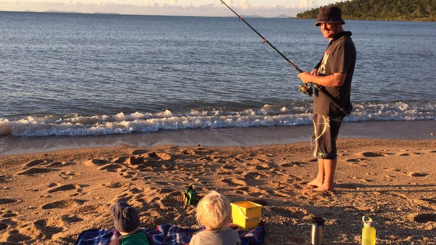 Grant McEwan smiles as he fishes on a beach, looking at his two children sitting on a blanket on the sand.