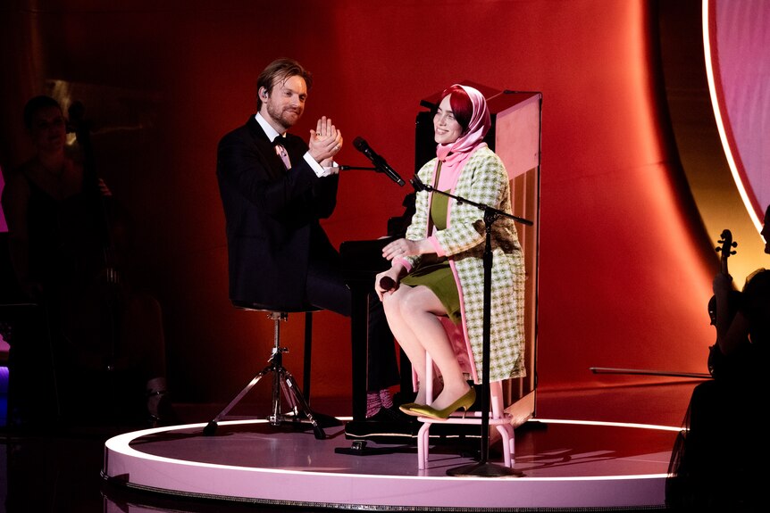 Finneas, sitting at a piano on stage, claps for Billie for smiles as she sits on a stool near him