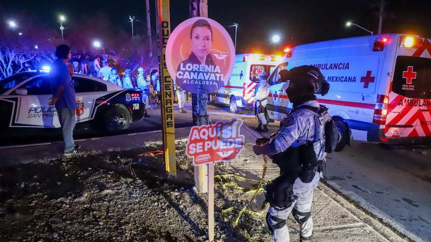 A man in military gear wraps yellow tape around a campaign sign as ambulances and police cars light the night behind him.