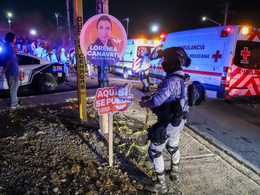 A man in military gear wraps yellow tape around a campaign sign as ambulances and police cars light the night behind him.