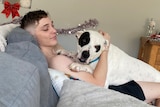 young man with short hair sits on couch whilst patting dog on his chest