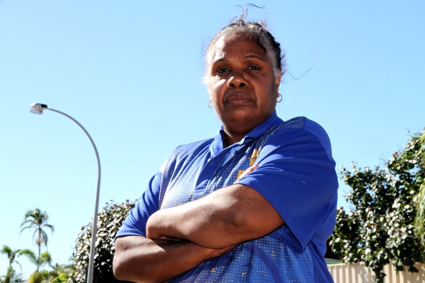 A woman wearing a purple t-shirt stands with her arms folded with a fence and street light in the backgorund.