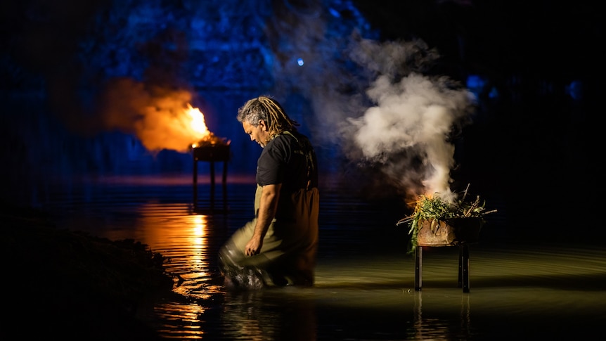 A man with dreadlocks wading into a river at night. Smoking eucalyptus leaves and fire in the background and mirrored in water