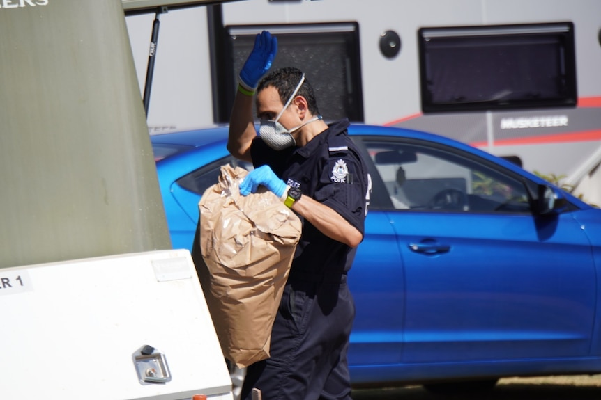 A police officer wearing a mask and gloves, carrying a brown paper bag.