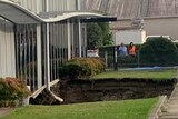 A hole in the ground in front of a building with two people looking over a fence