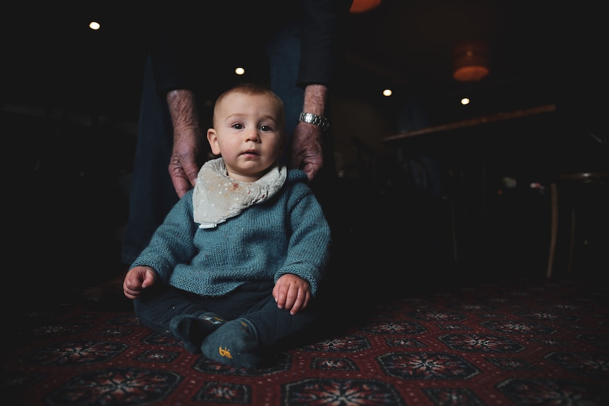 A baby boy wearing a knitted jumper and bib sits on a red carpeted floor.