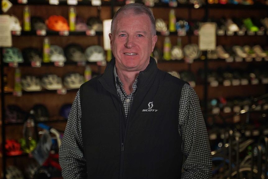 A man in a checked shirt and black vest stands in front of bicycle helmets in a shop.